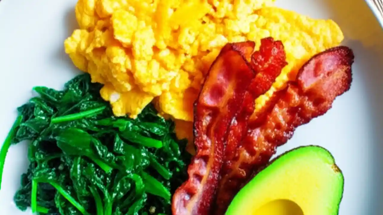 A visually appealing, healthy ketogenic breakfast featuring scrambled eggs, sliced avocado, crispy bacon, and fresh spinach on a rustic wooden table.